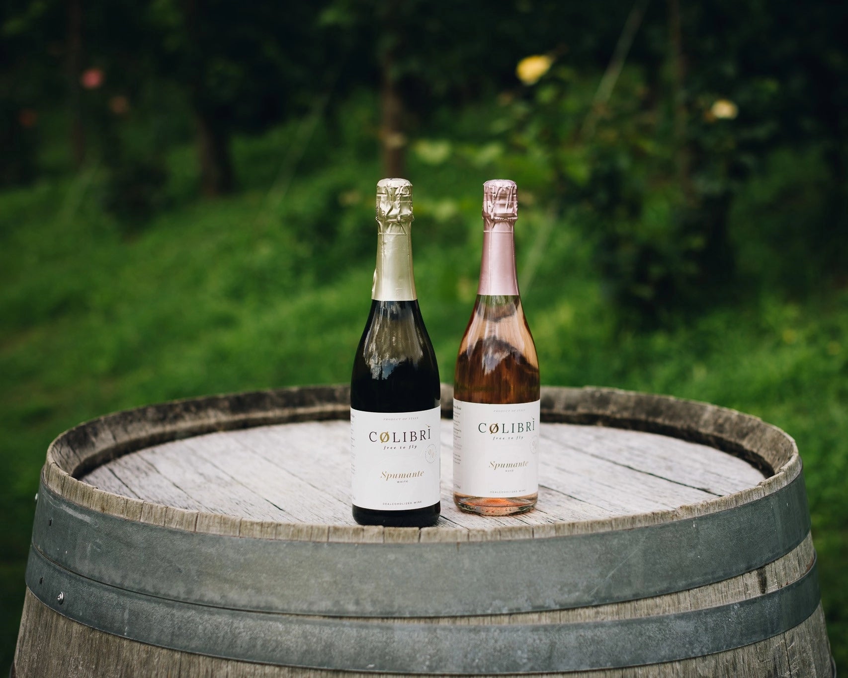 Colibri alcohol-free sparkling wine rosé and white bottles displayed on a wooden barrel in a green vineyard landscape.