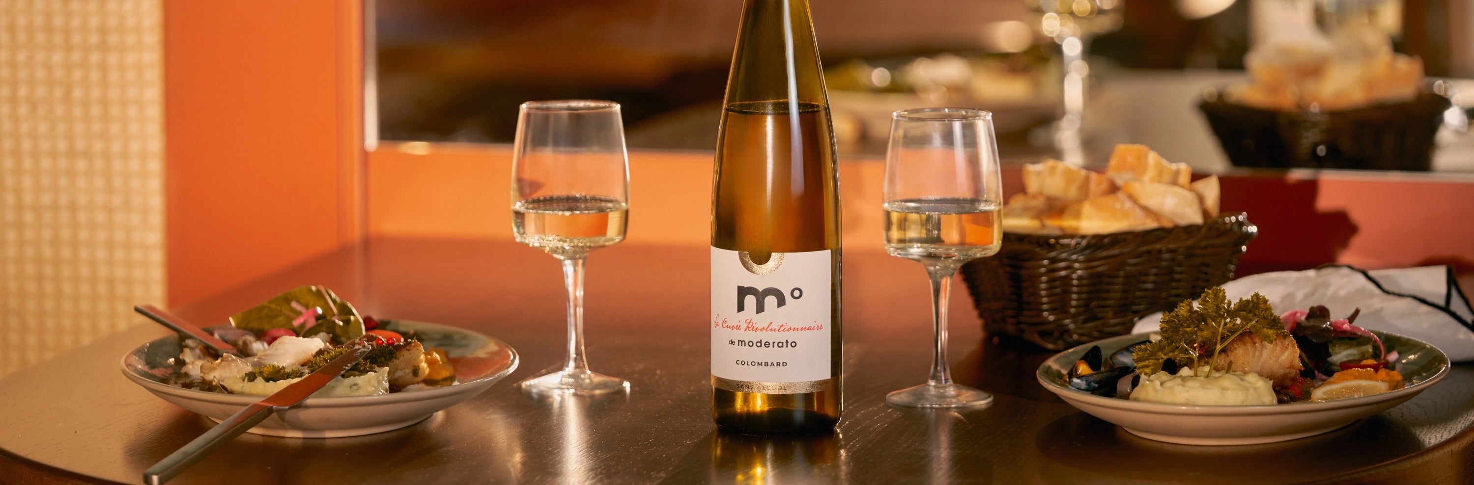 Moderato Colombard alcohol-free white wine served with two glasses and plated dishes in a warm restaurant setting.