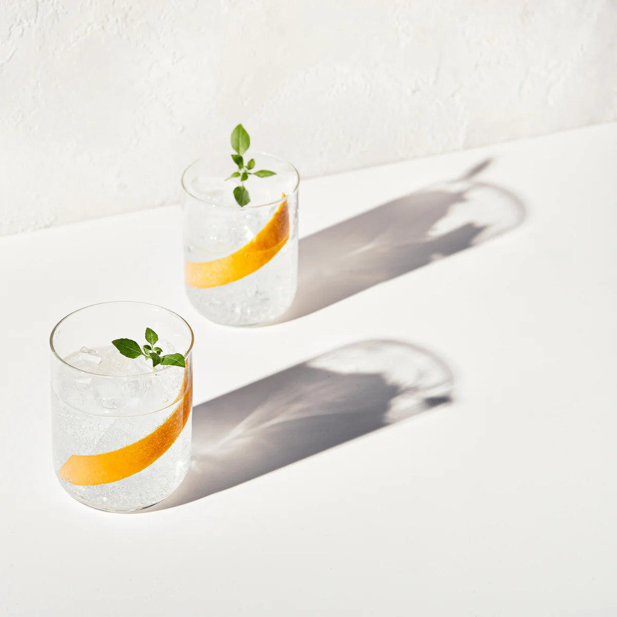 Two glasses of seved Nona June and Tonic mocktail with orange zest and mint leaves on a white surface