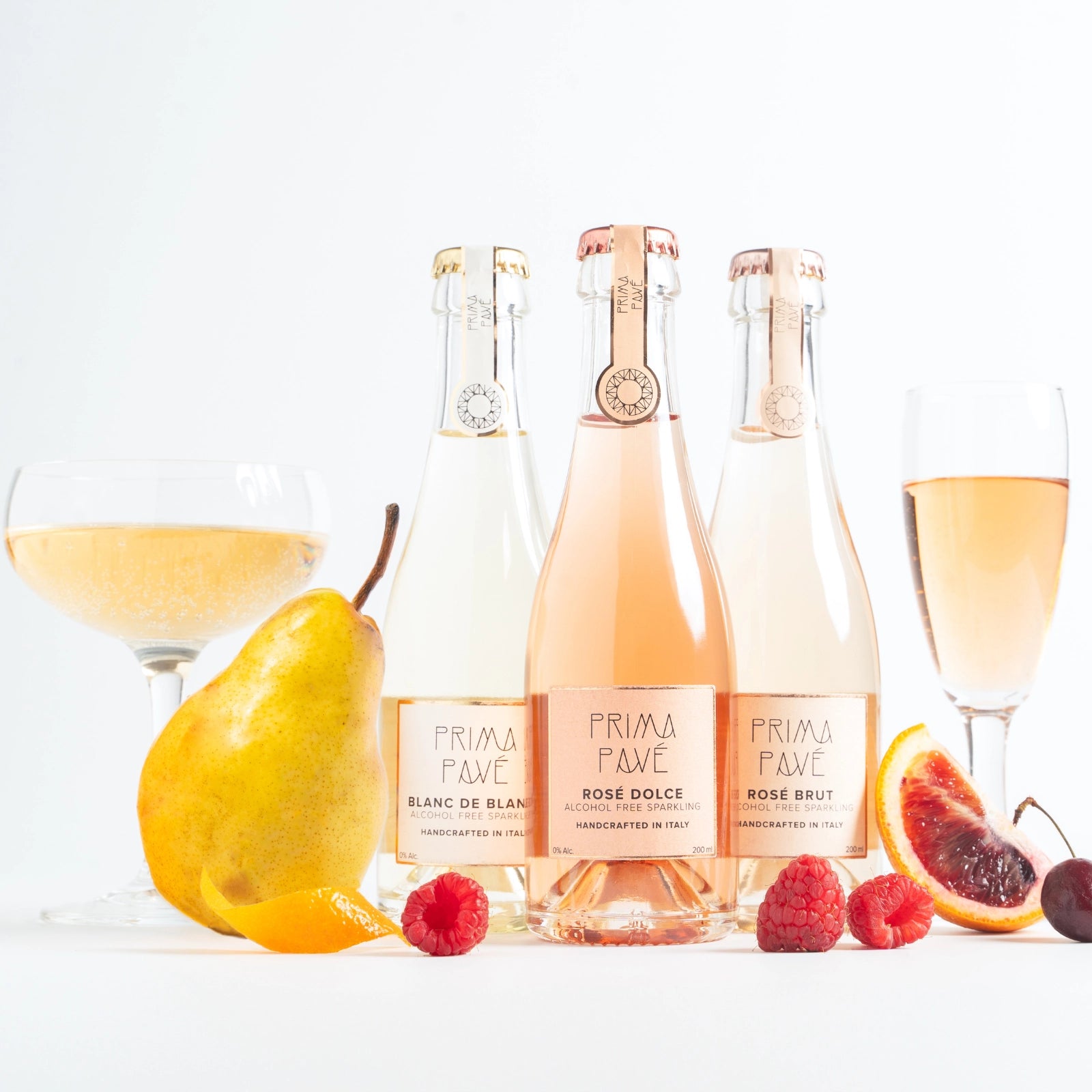 Three bottles of Prima Pavé alcohol-free sparkling wine with glasses and fresh fruits such as pear, raspberries, and citrus arranged in a bright lifestyle scene.