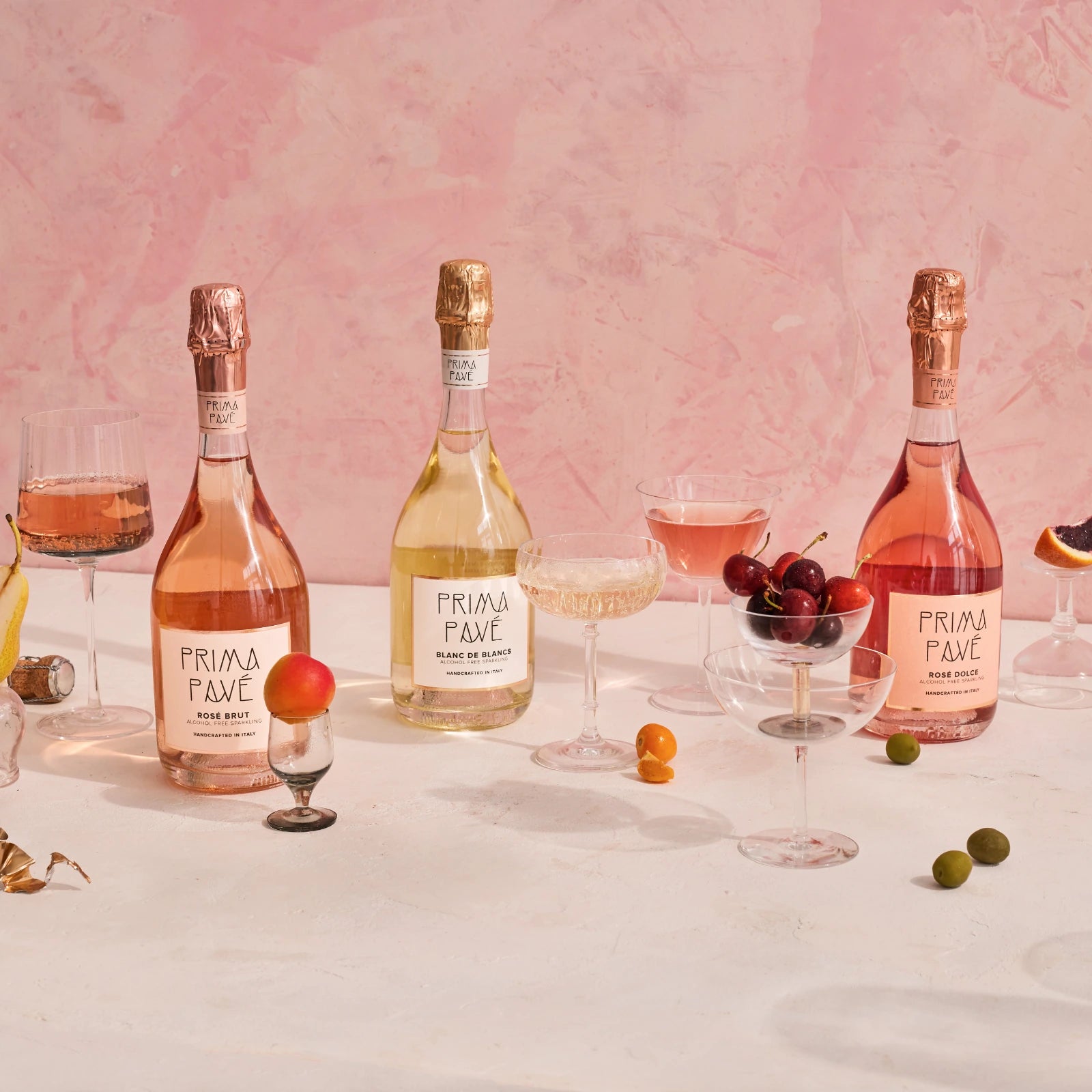 Prima Pavé alcohol-free sparkling wines — Rosé Brut, Blanc de Blancs, and Rosé Dolce — styled with glasses, fruits, and decorations against a pink artistic backdrop.