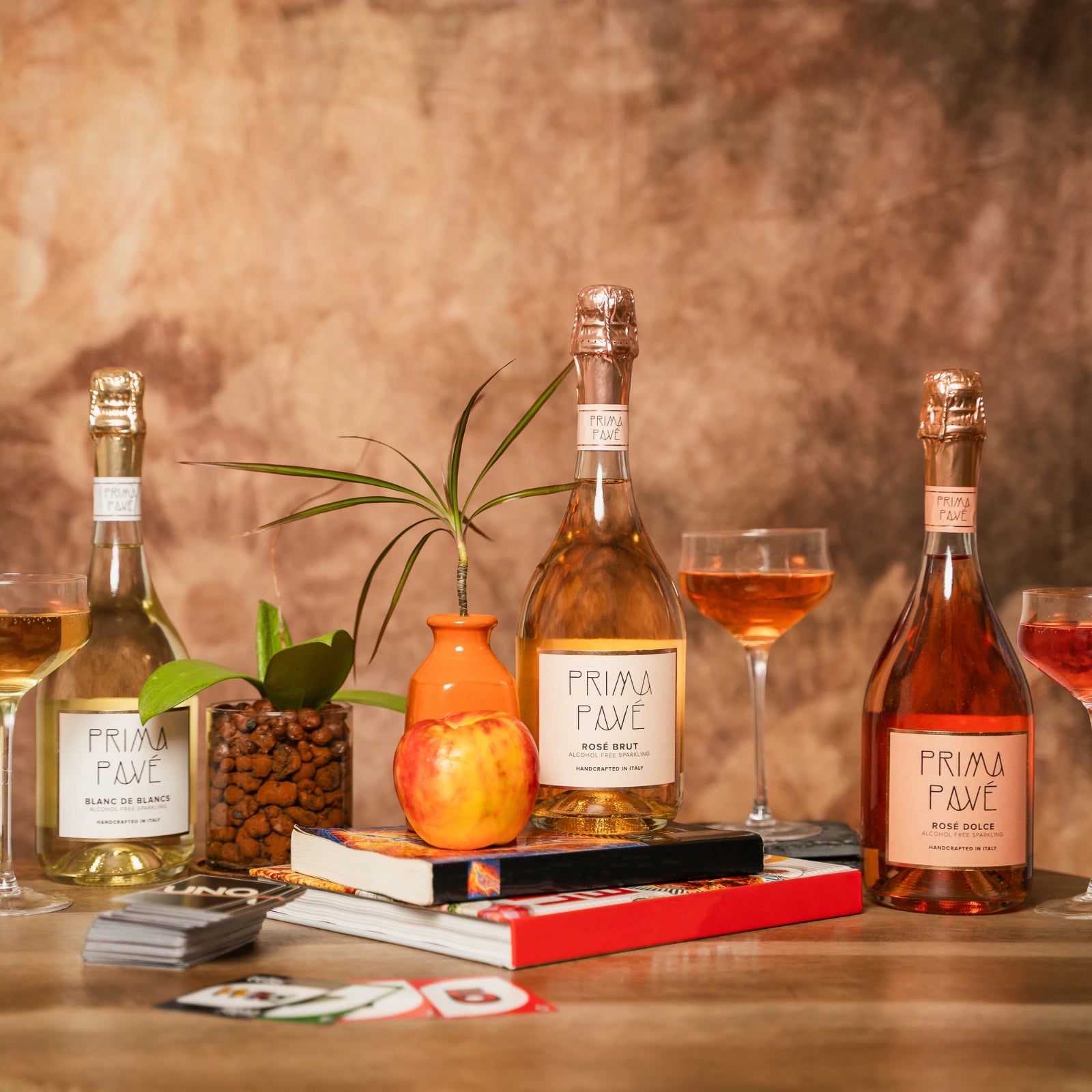 Prima Pavé alcohol-free sparkling wines displayed in a cozy lifestyle scene with books, plants, and cocktails in warm earthy tones.