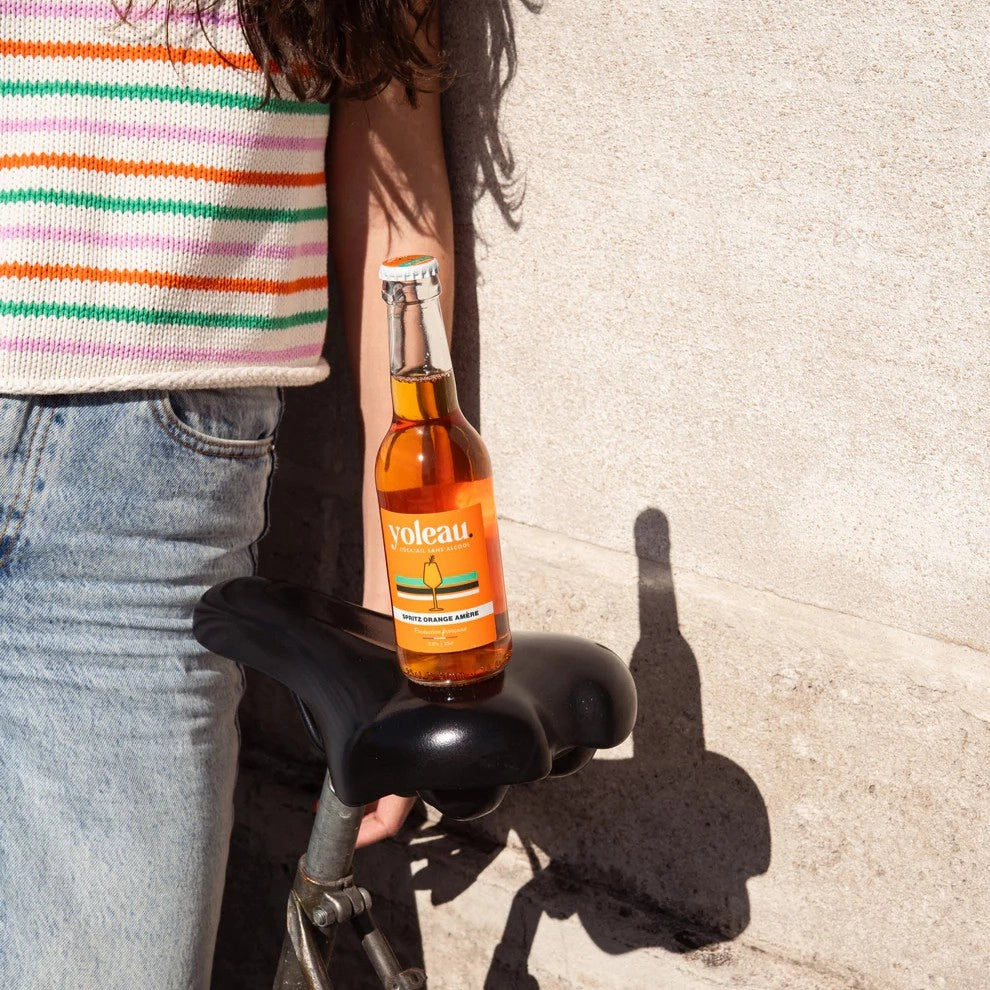 Yoleau alcohol-free orange spritz bottle placed on a bicycle seat in a sunny urban setting.