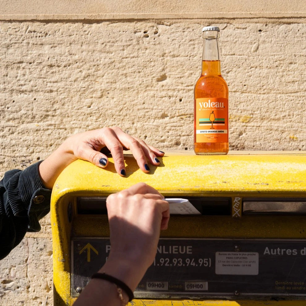 Yoleau alcohol-free orange spritz bottle on a yellow mailbox with a person posting a letter in a sunny street scene.
