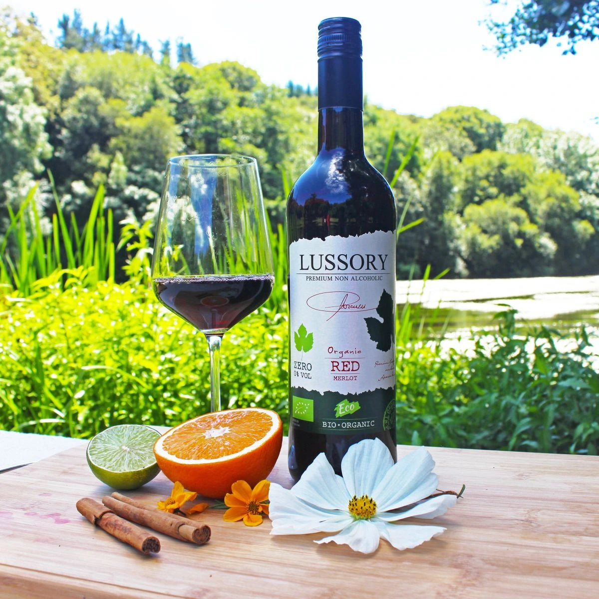 LUSSORY Organic Merlot Non-Alcoholic Red Wine bottle with a poured glass outdoors, surrounded by orange, lime, cinnamon sticks, and flowers on a wooden table.