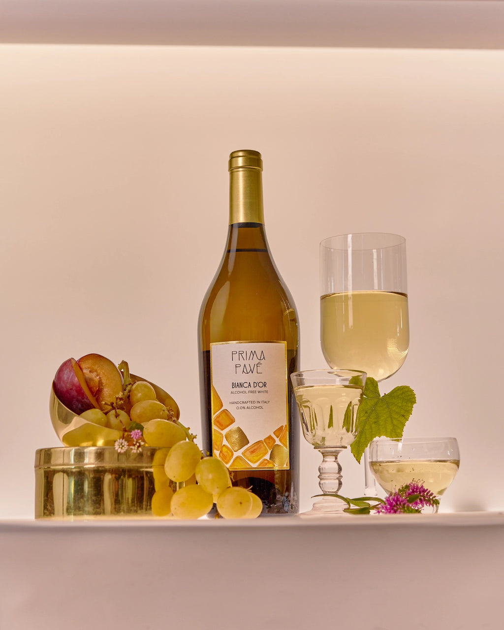 Láhev 750 ml nealkoholického bílého vína PRIMA PAVÉ Bianca D’Or se sklenicemi, čerstvými hrozny, švestkou a zlatými doplňky v elegantním, jemně osvětleném prostředí.