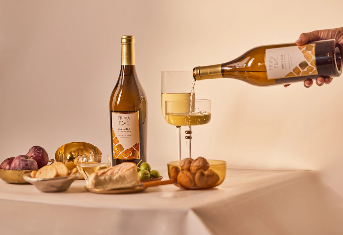 PRIMA PAVÉ Bianca D’Or Non-Alcoholic being poured into a glass, served with cheese, grapes, and walnuts on a light table setting.