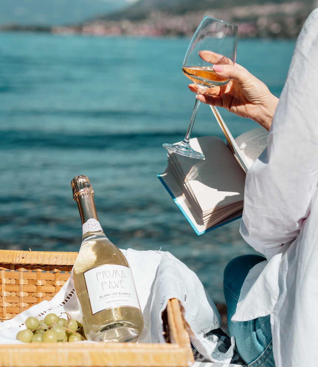 Person enjoying a glass of PRIMA PAVÉ Blanc de Blancs non-alcoholic sparkling wine while reading by the lake, with a picnic basket and grapes nearby.