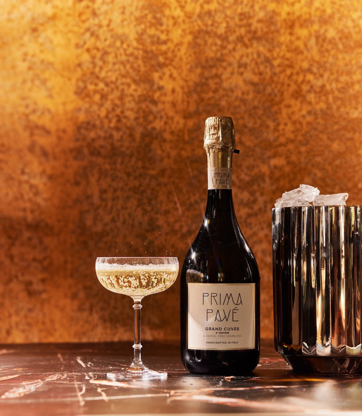 PRIMA PAVÉ Grand Cuvée Non-Alcoholic Sparkling Wine 750 ml bottle with a coupe glass of golden bubbles and ice bucket on an elegant marble table.