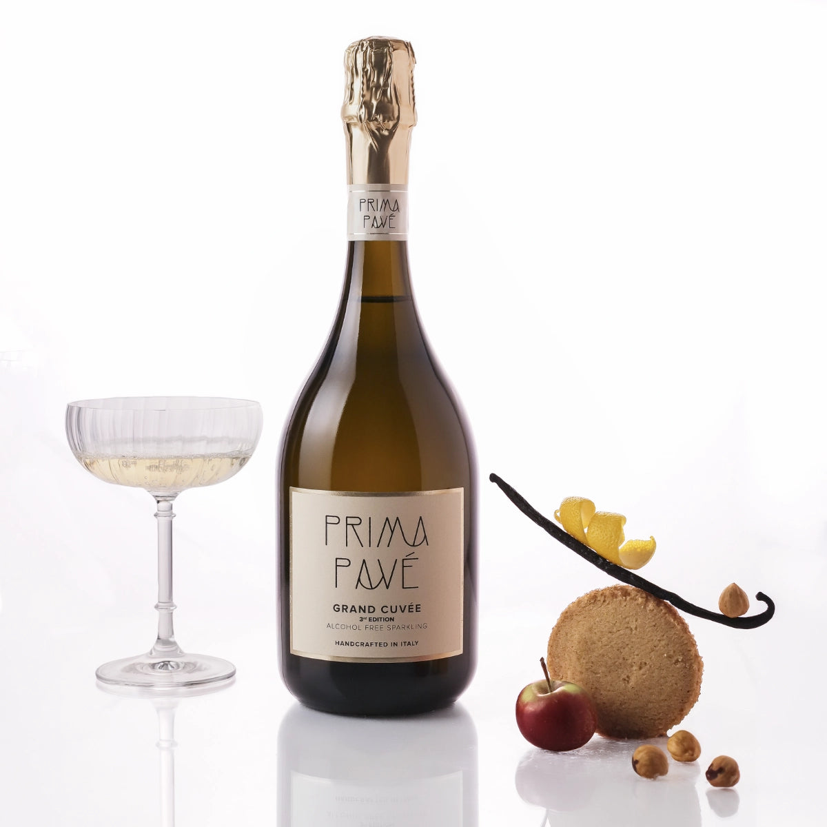 750ml bottle of PRIMA PAVÉ Grand Cuvée Non-Alcoholic Sparkling Wine with a filled coupe glass and cookies and nuts with fruits on a white  background.