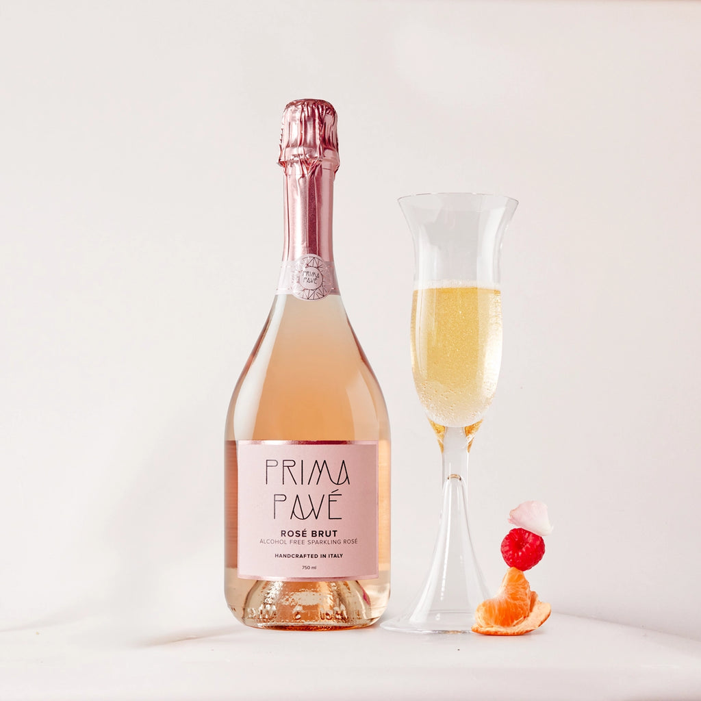 PRIMA PAVÉ Rosé Brut Non-Alcoholic Sparkling Wine bottle with filled glass and fresh fruit garnish on white background
