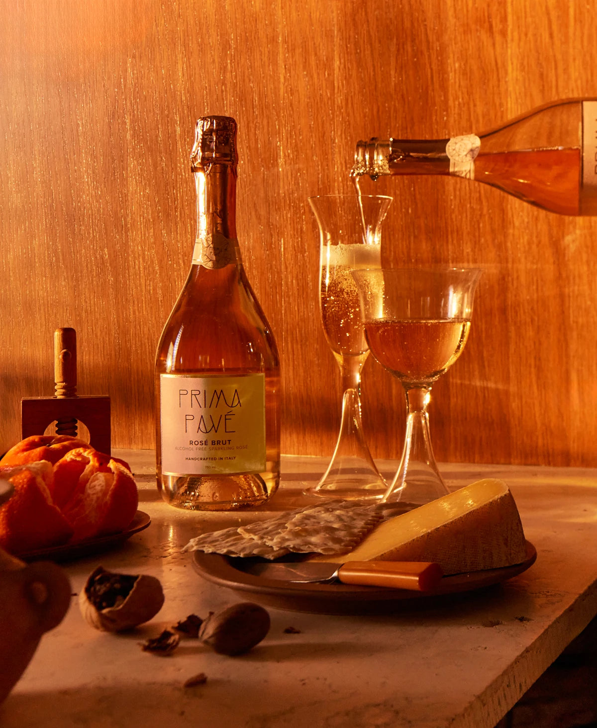 PRIMA PAVÉ Rosé Brut Non-Alcoholic Sparkling Wine served with cheese, citrus, and nuts in warm golden light, showcasing an elegant pairing setup