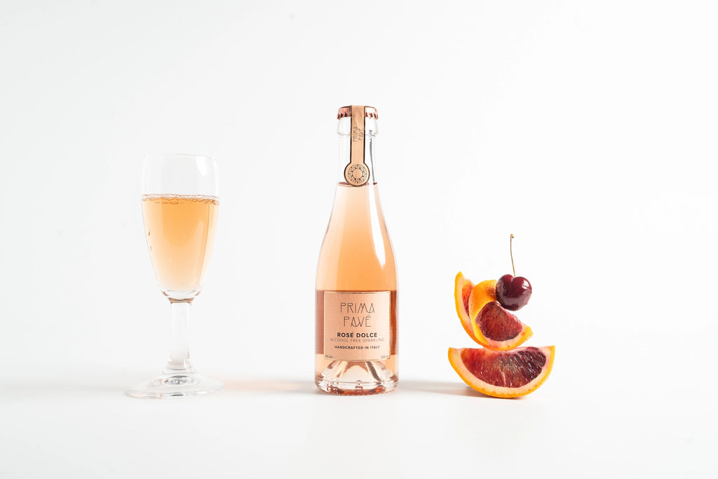 Bottle of PRIMA PAVÉ Non-Alcoholic Sparkling Wine with a filled glass and sliced fruit on a white background