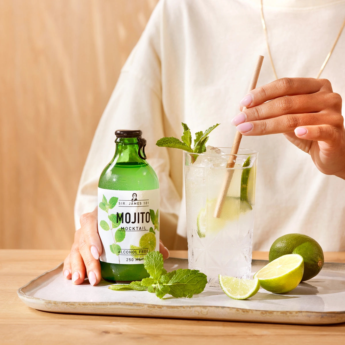 A person holding a straw in a glass of SIR. JAMES 101 Mojito Alcohol Free mocktail with lime and mint on a wooden table.