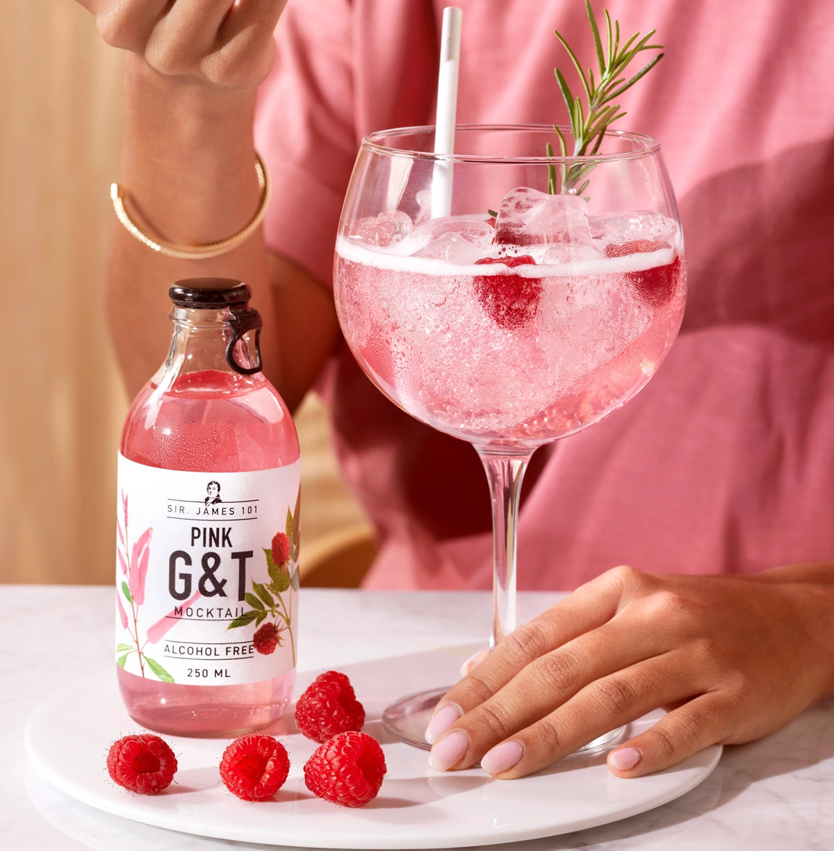 Person holding a glass of SIR. JAMES 101 Pink G&T Alcohol Free mocktail with raspberries and ice, next to the bottle on a white plate